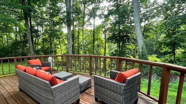 Une terrasse en bois, dotée de meubles en osier et d’une rambarde, entourée d’arbres.