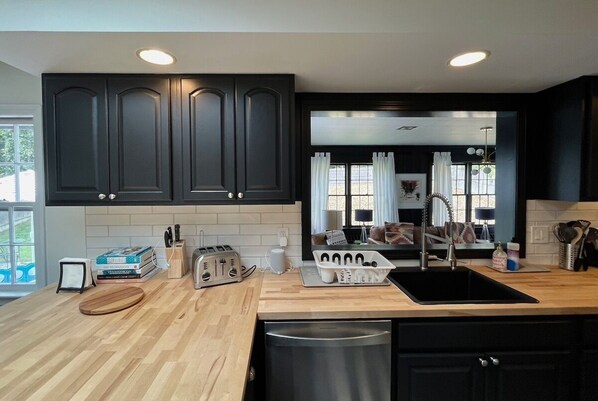 A modern kitchen with dark cabinets, a wooden countertop, a sink, and a view into a living area.
