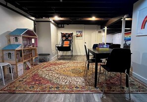A room with a playhouse, a basketball hoop, a dining table with chairs, and a colorful rug.