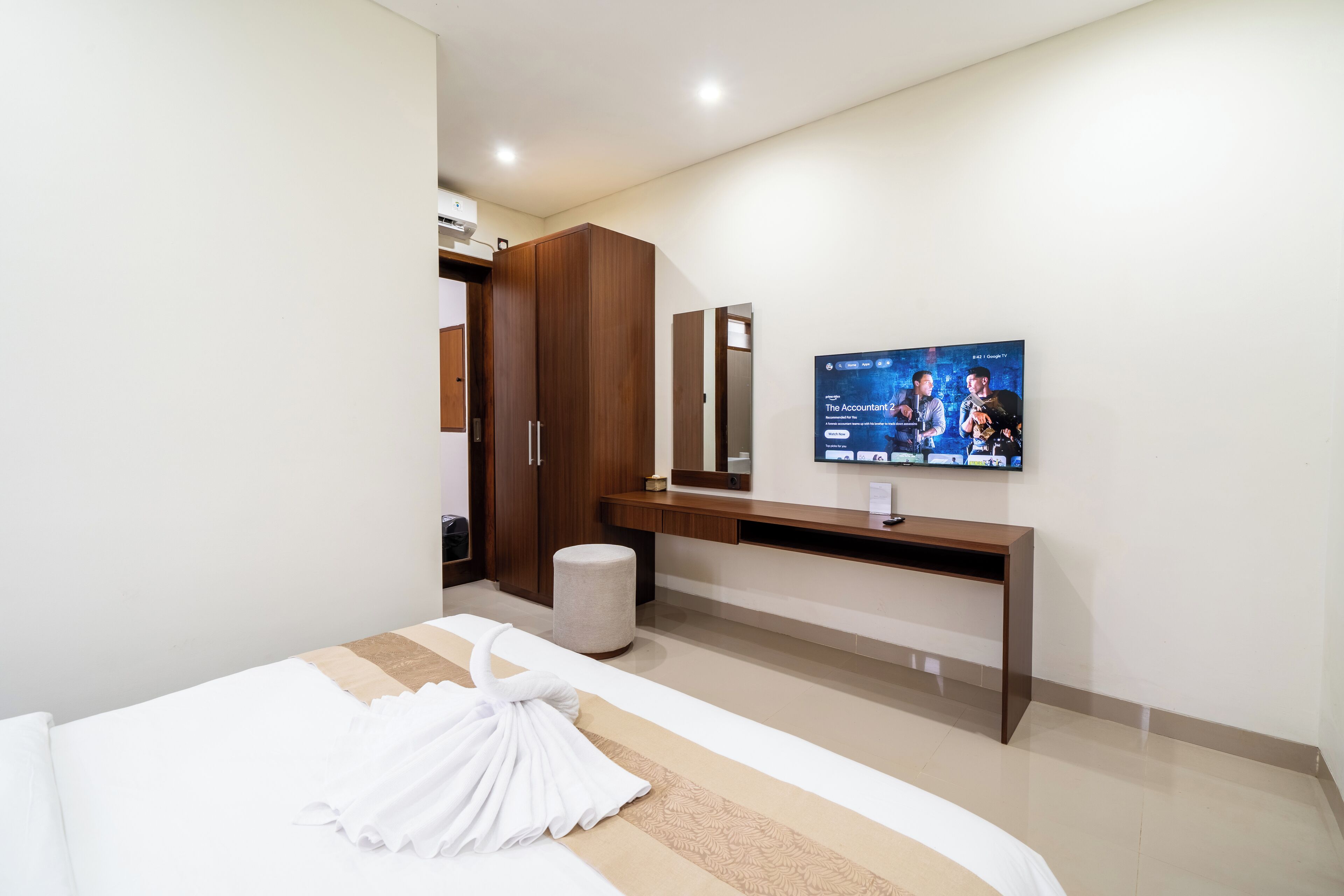 Habitación de hotel con cama, televisor montado en la pared, armario de madera y un taburete pequeño.