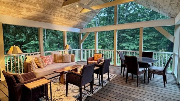 Eine überdachte Terrasse im Freien mit Holzboden, einer Couch, Stühlen, einem Tisch und einer Geländer.
