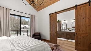 Un dormitorio con una cama grande, techo de madera, puerta corredera al baño y vistas a árboles.