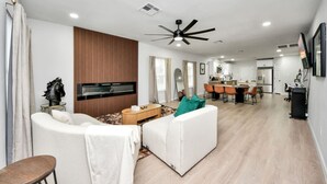 Ein modernes Wohnzimmer mit Flachbildfernseher, Deckenventilator und Essbereich. Im Hintergrund ist eine Küche zu sehen.