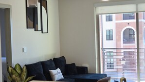 Un soggiorno moderno con un divano, un tavolo da pranzo con sedie, una ventola a soffitto e vista su un edificio in mattoni attraverso una grande finestra.