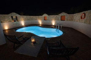 A swimming pool with a curved edge, surrounded by stone walls and a tiled deck. There are two lounge chairs and a table with a lantern. - Villa Joy Mare - Private Pool Parking in Chania (Chania)