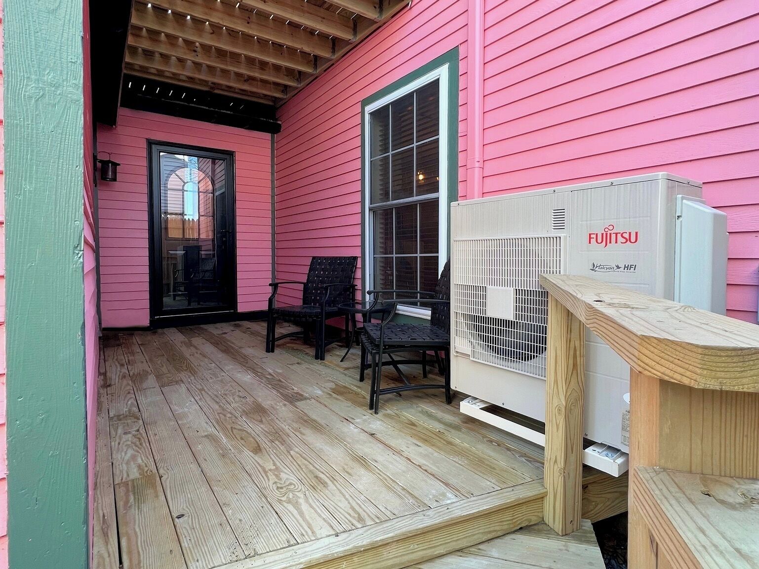 A pink house with a wooden deck, two black chairs, and a white air conditioning unit.