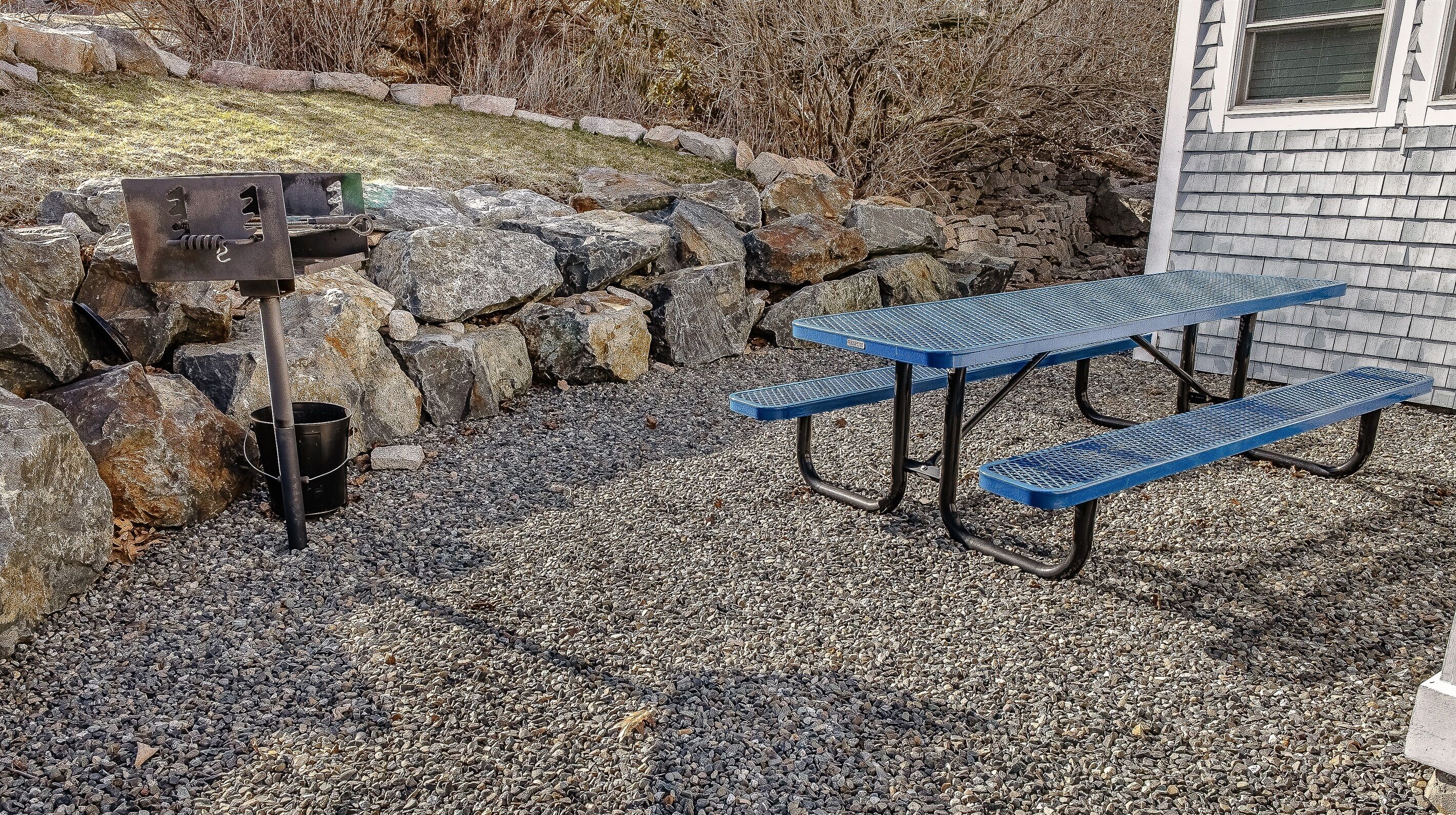 A picnic table with benches, a metal grill, and a stone wall.