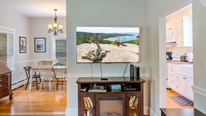 A living room with a television displaying a scenic view, a dining area with a table and chairs, and a kitchen with white cabinets. - Acadia View Cottage: Peaceful, Pet Friendly, Fire Pit, Near Peaks and Echo Lake (Mount Desert)