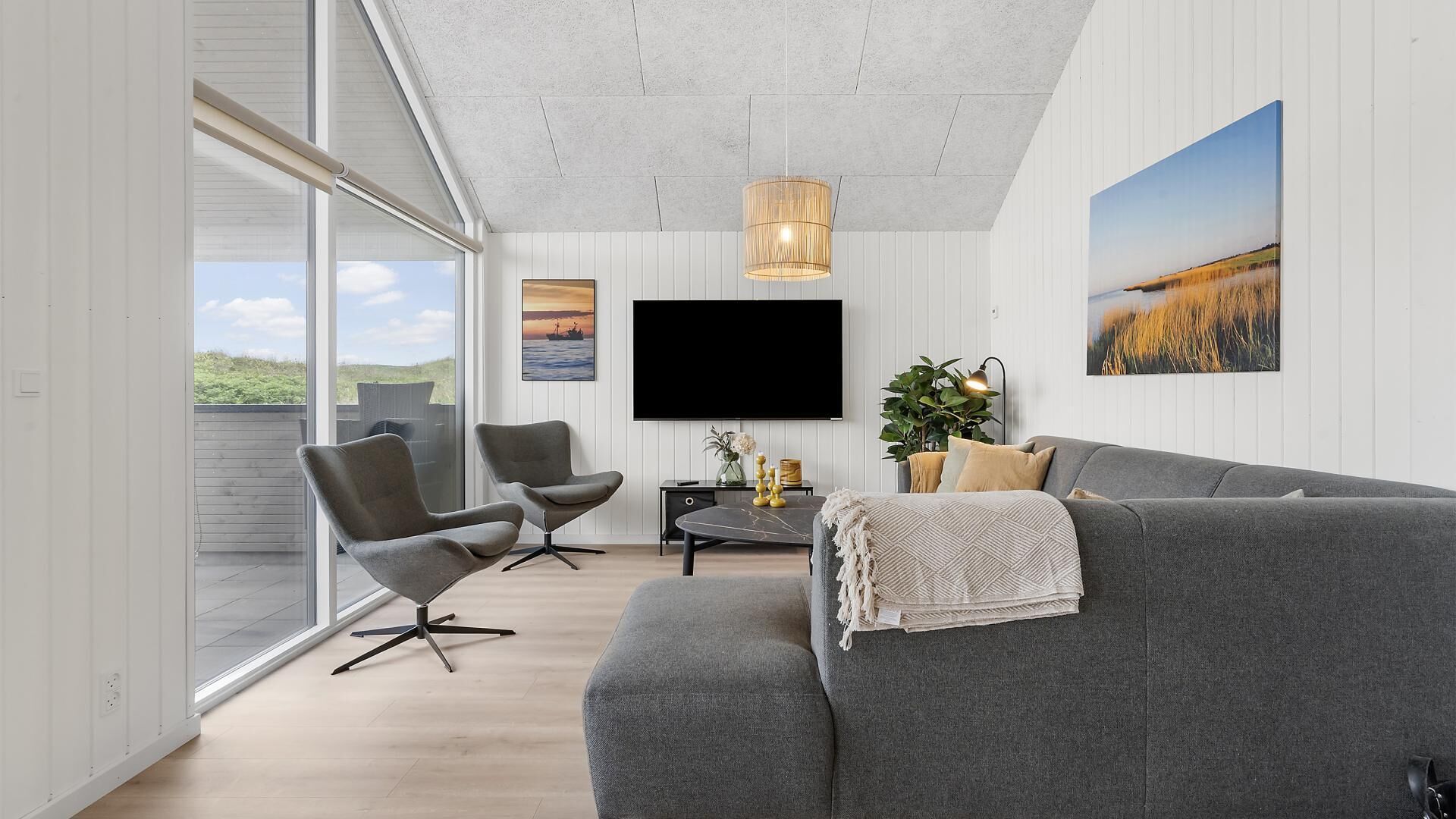 En moderne stue med en grå sofa, to lænestole, et fladskærms-tv og et stort vindue med udsigt over landskabet.