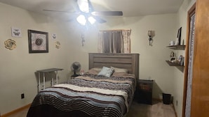 Une chambre à coucher avec un lit, un ventilateur de plafond, une table de chevet et une commode, ainsi qu’une porte.