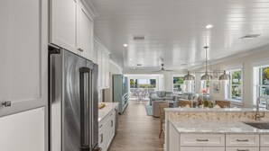 A modern kitchen with white cabinetry, a stainless steel refrigerator, a marble island with a sink, and a dining area with a view of the outdoors. - A Perfect Day: Gulf Front! Rooftop Deck, Putt Putt, Pool Spa by Anna Maria Life (Anna Maria)