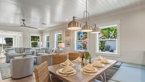 A spacious living room with a large dining table set for a meal, a ceiling fan, and large windows offering a view of the outdoors. - A Perfect Day: Gulf Front! Rooftop Deck, Putt Putt, Pool Spa by Anna Maria Life (Anna Maria)