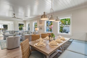 A spacious living room with a large dining table set for a meal, a ceiling fan, and large windows offering a view of the outdoors. - A Perfect Day: Gulf Front! Rooftop Deck, Putt Putt, Pool Spa by Anna Maria Life (Anna Maria)
