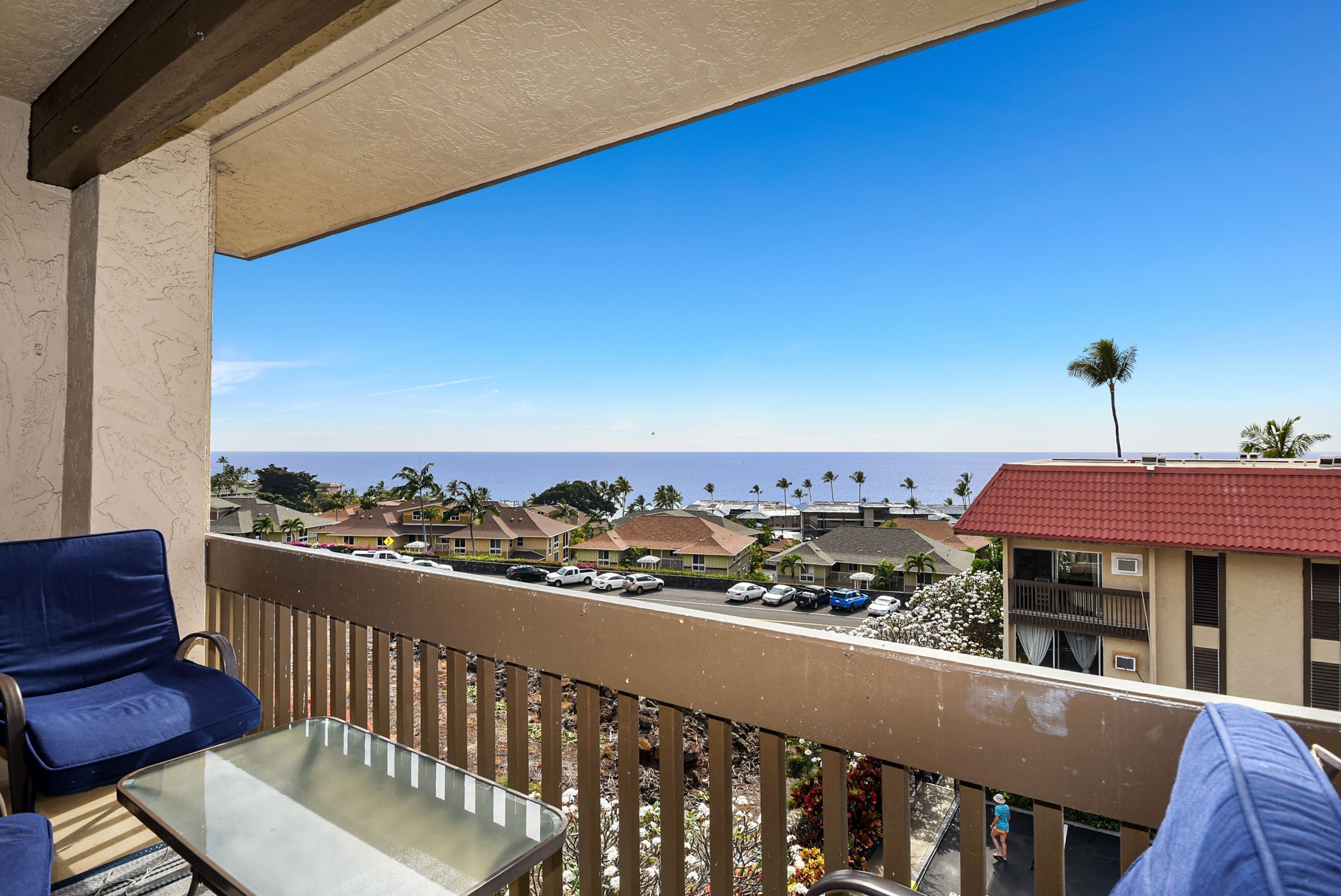 A balcony with a glass table and blue chairs, overlooking a street with parked cars and buildings, and a clear blue sky.