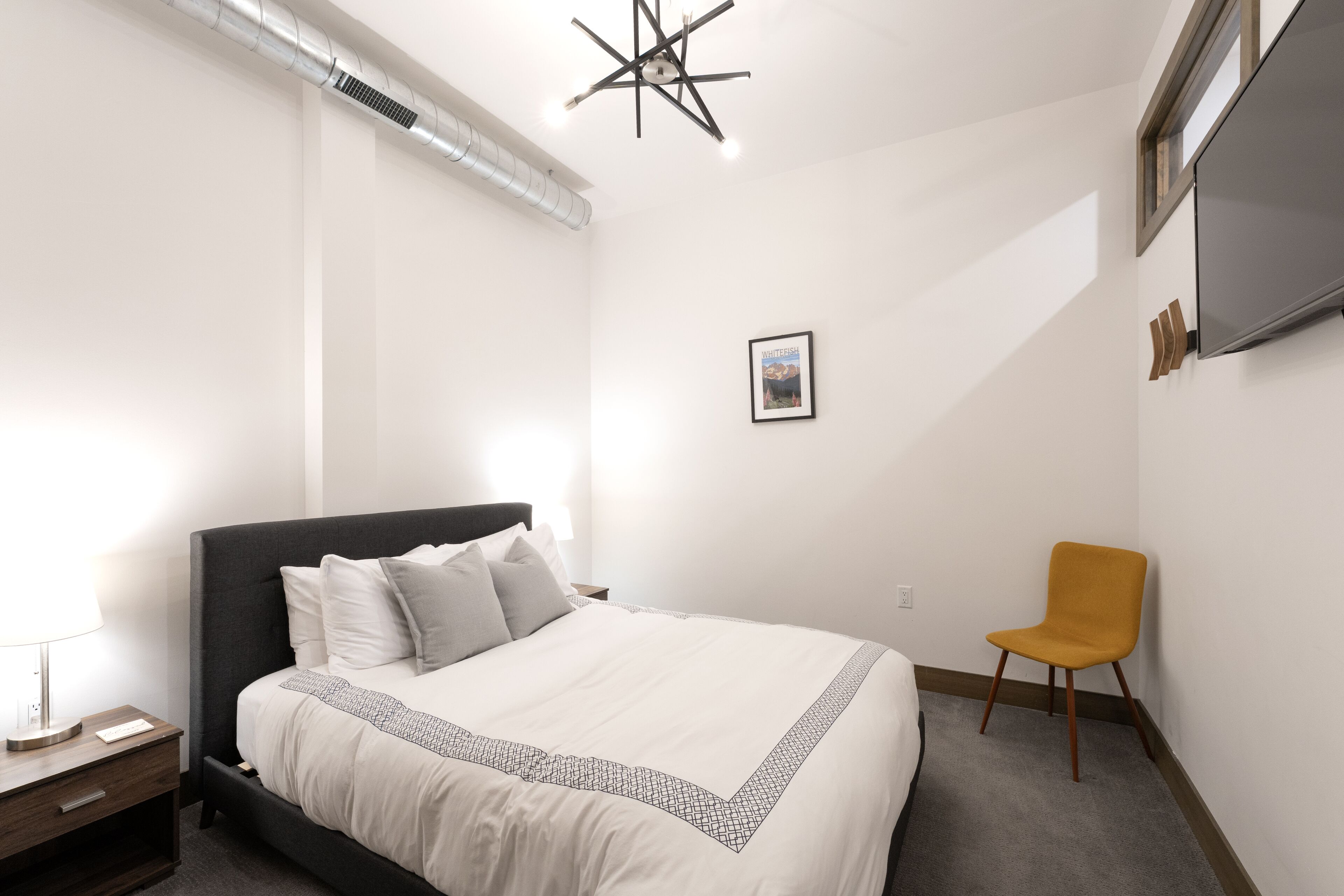 A bedroom with a bed, a nightstand, a chair, a ceiling light fixture, and a wall-mounted TV.