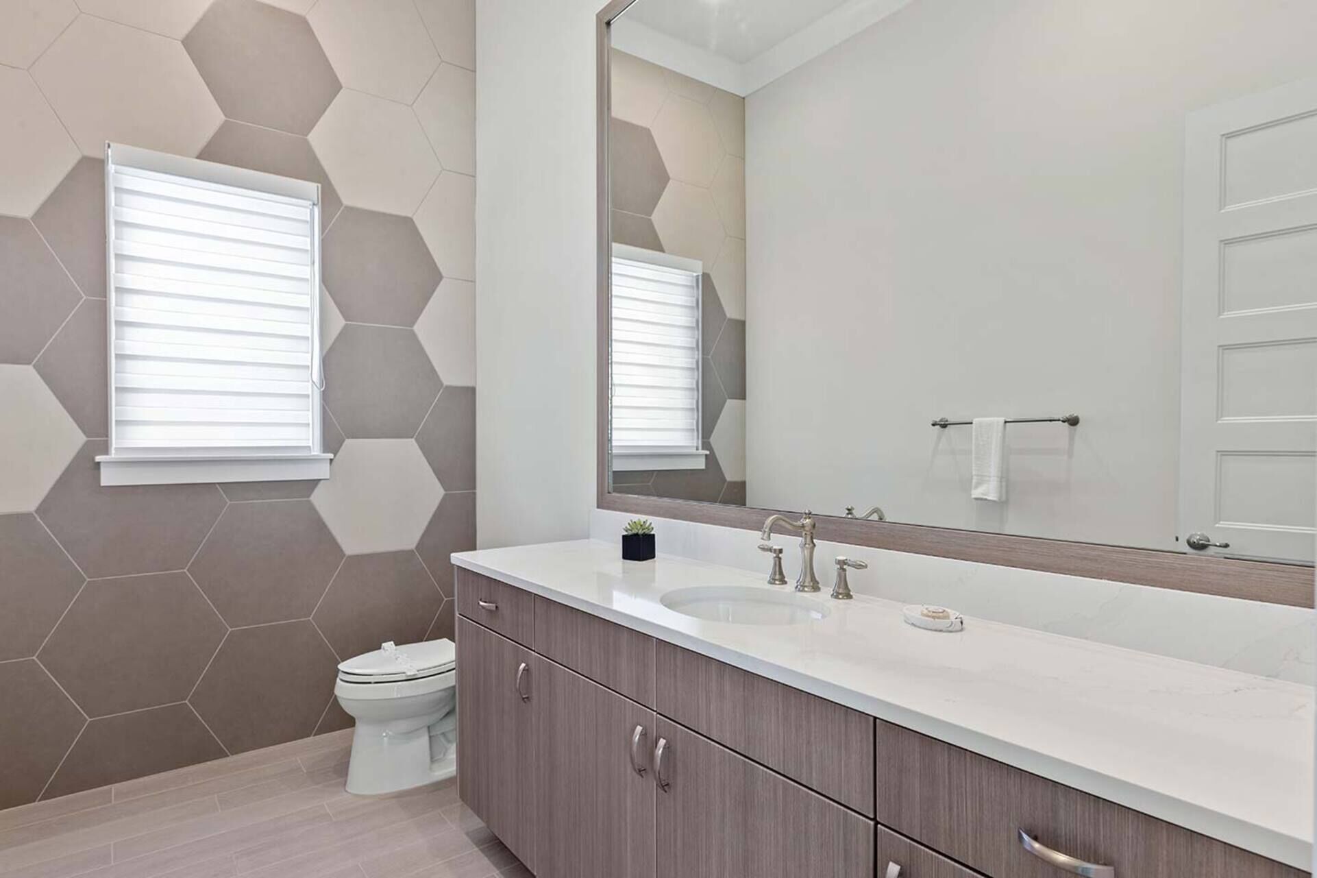 Un baño moderno con pared de azulejos hexagonales, un espejo grande, un lavabo blanco y un inodoro.