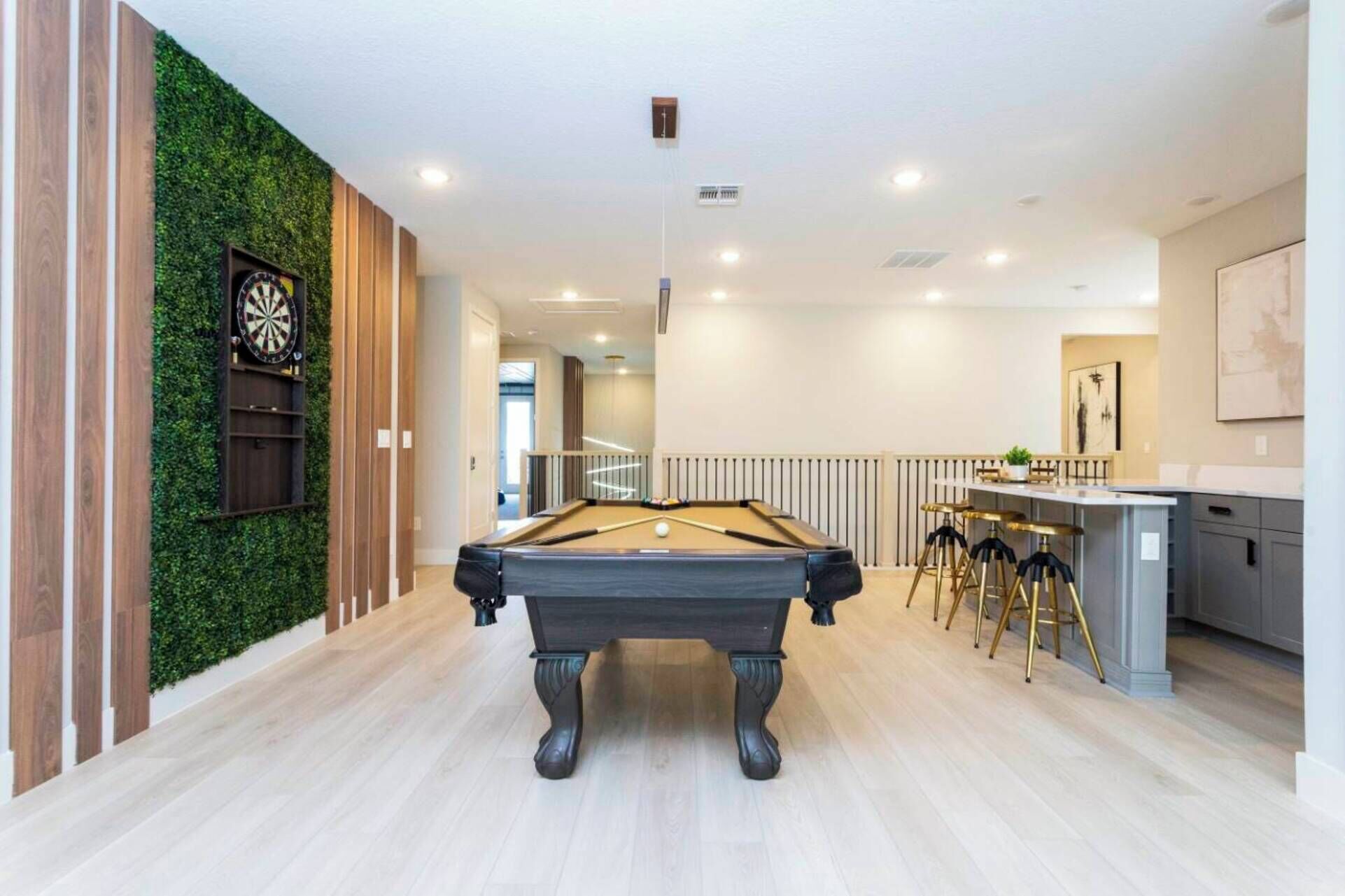 A pool table in a modern room with a green wall, a bar area with stools, and a clock on the wall.