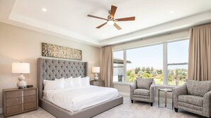 A hotel room with a large bed, two bedside tables with lamps, a ceiling fan, a view of the outdoors, and a seating area with two chairs. - 5 Star Villa Aurelius – A Golden Grand Residence (Orlando)