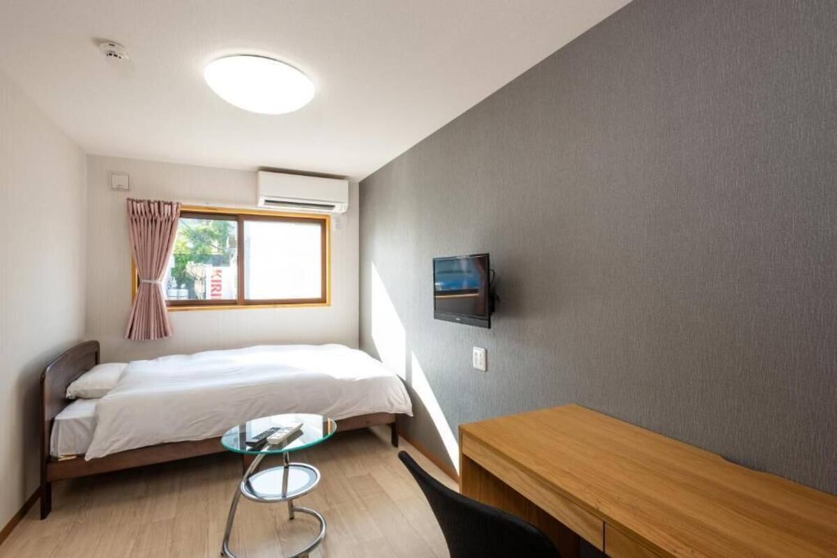 A small, well-lit room with a bed, a TV mounted on the wall, and a wooden desk.