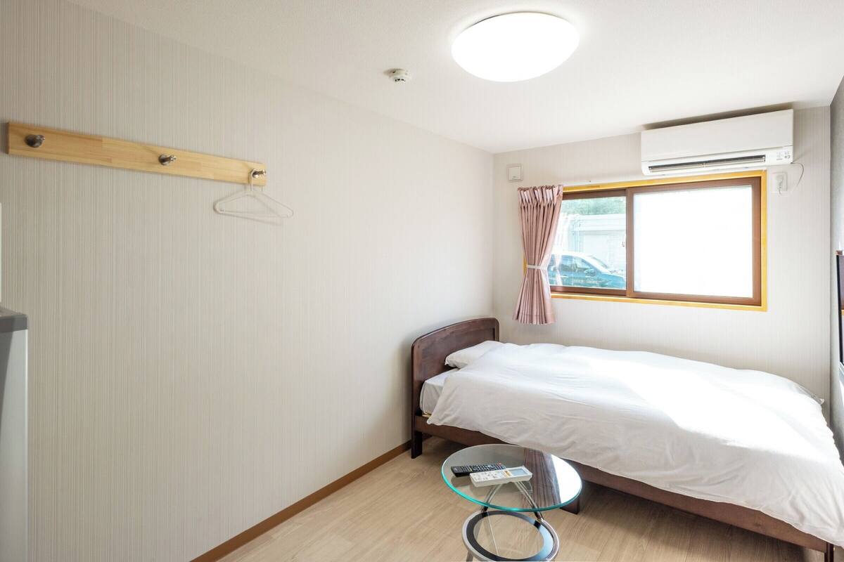 A small, neatly arranged room with a single bed, a bedside table with a remote control, a hanging coat rack, a window with curtains, and an air conditioning unit.