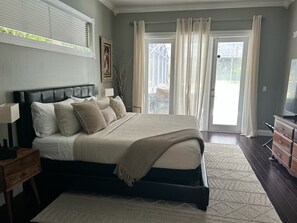 Ein Schlafzimmer mit Bett, Nachttisch, Kommode, Fernseher, Fenster mit Jalousien und einer Tür mit Vorhängen.