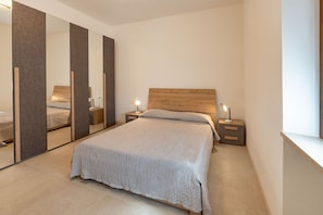 A bedroom with a large mirror, a wooden headboard, a bed with a grey bedspread, two bedside tables with lamps, and a window.