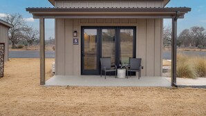 A building with a number '5' sign, a covered patio area with two chairs and a table, and a view of a road and trees in the background. - Silverstone Casita 5 at Featherstone Ranch (Stonewall)