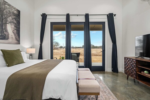 Un dormitorio con una cama grande, un banco, un televisor y vistas a un campo por la ventana.
