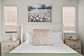 Une chambre à coucher avec un grand lit, deux tables de chevet, un tableau au-dessus du lit et une fenêtre avec un store.
