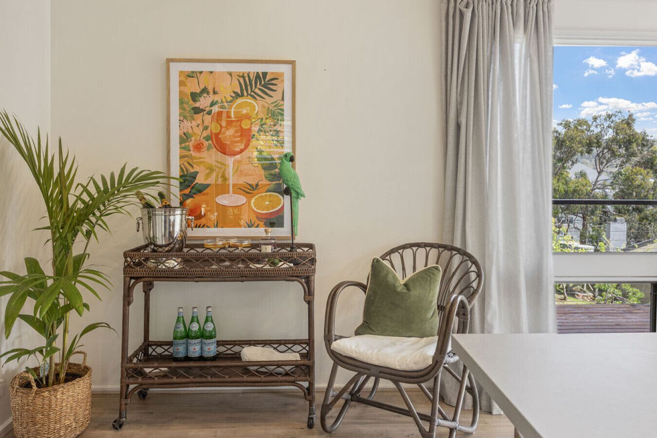 Une pièce avec une table en osier sur laquelle sont posées des bouteilles, une chaise verte, un tableau représentant une boisson et une vue sur des arbres par la fenêtre.
