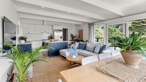 A modern kitchen with white cabinets, a blue island, and a dining area with a wooden table and blue sofa. - NEW! The Soak House- Walk to the beach! (Lorne)