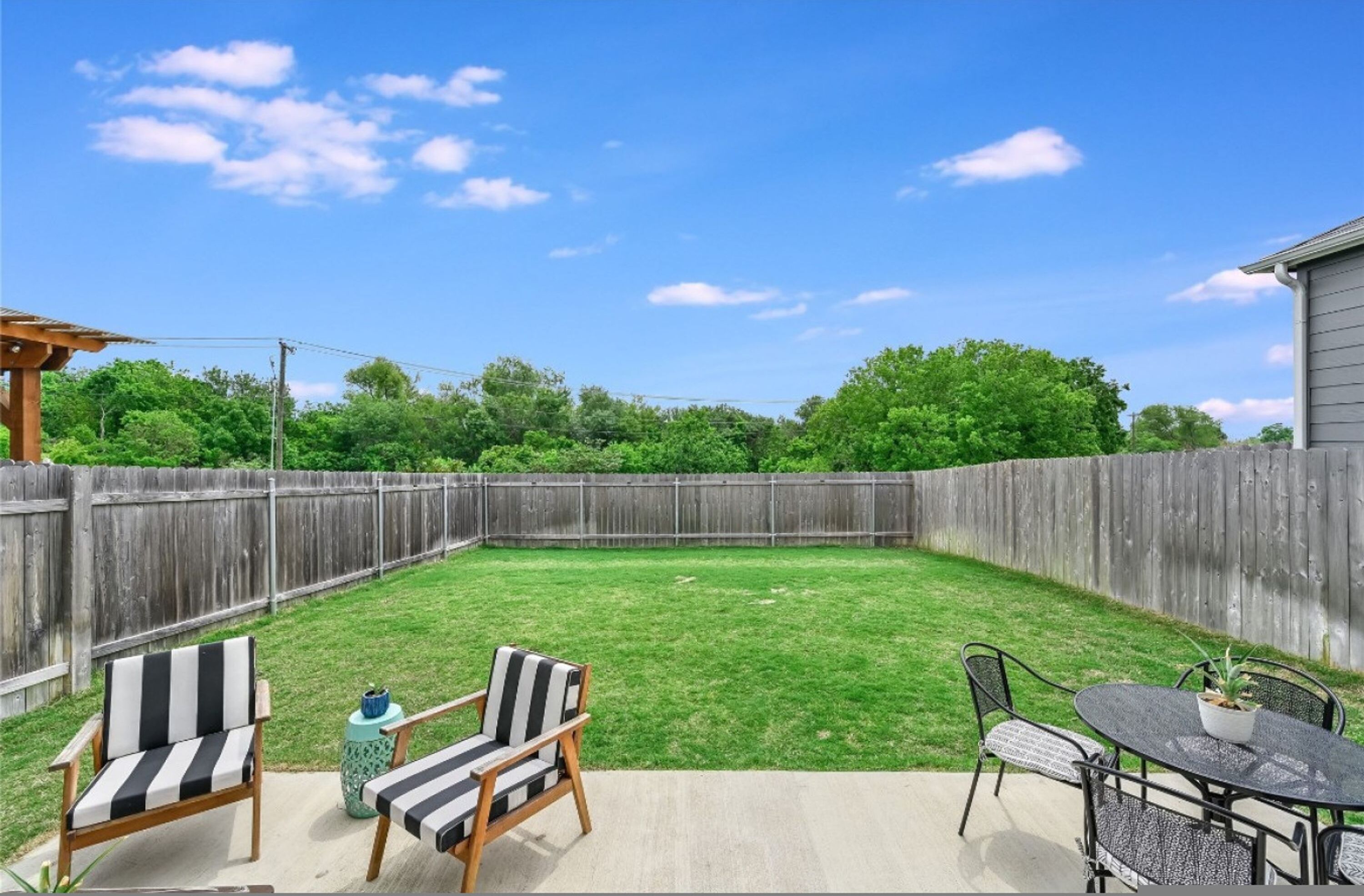A backyard with a wooden fence, a concrete patio, and outdoor furniture including two striped chairs and a table.