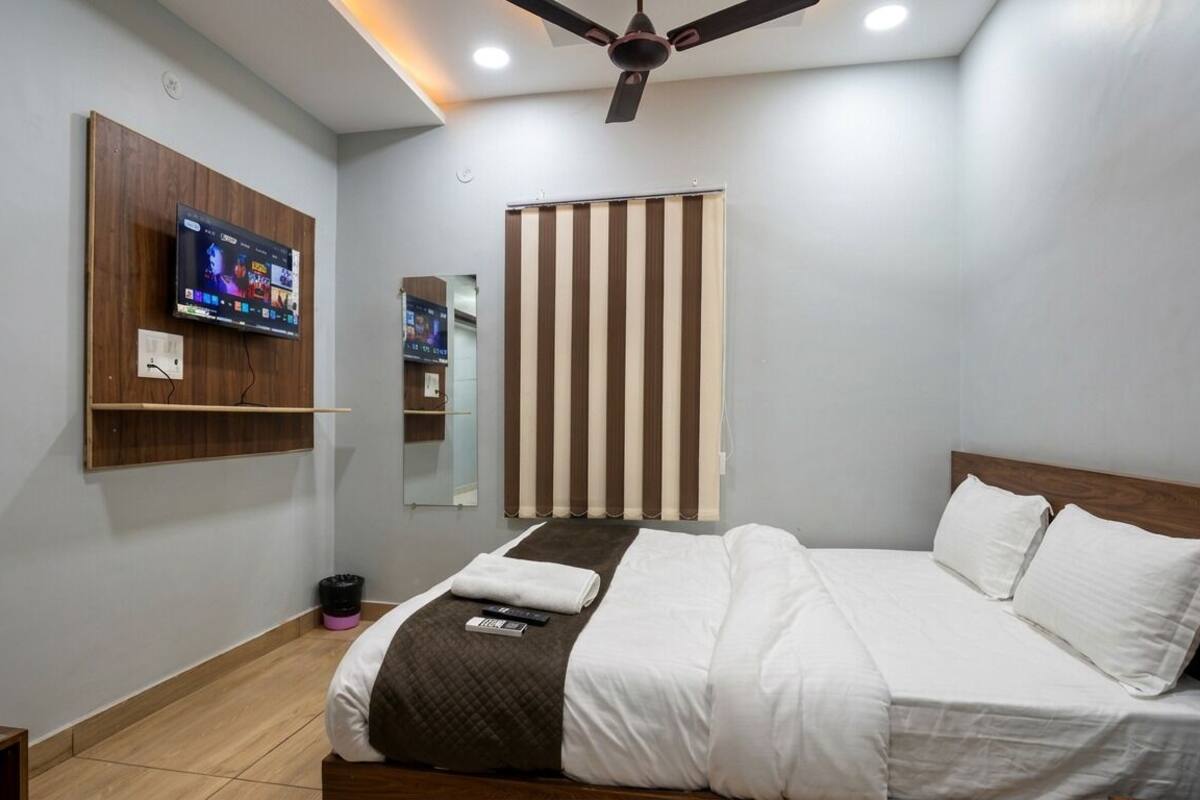 A modern hotel room with a bed, a TV mounted on the wall, a ceiling fan, and a window with vertical blinds.