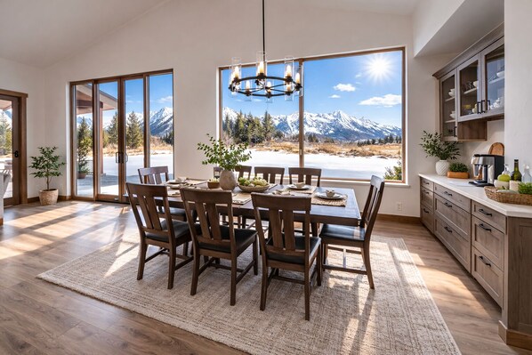A dining area with a large table and chairs, a chandelier, and a view of snow-capped mountains through large windows. - Brand New Luxury Estate by Jackson Hole Rentals™ (Jackson)