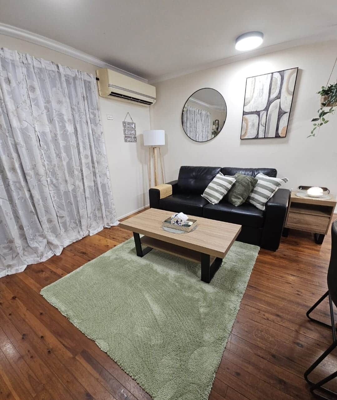 A living room with a wooden coffee table, a black sofa with cushions, a round mirror, and a painting on the wall.