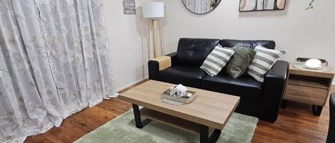 A living room with a wooden coffee table, a black sofa with cushions, a round mirror, and a painting on the wall.