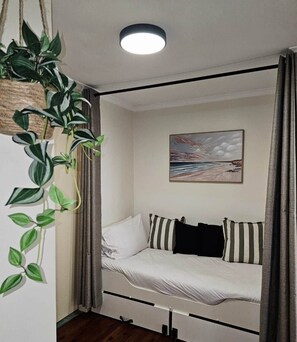 A small, cozy room with a bed, a ceiling light, a painting, and a plant.
