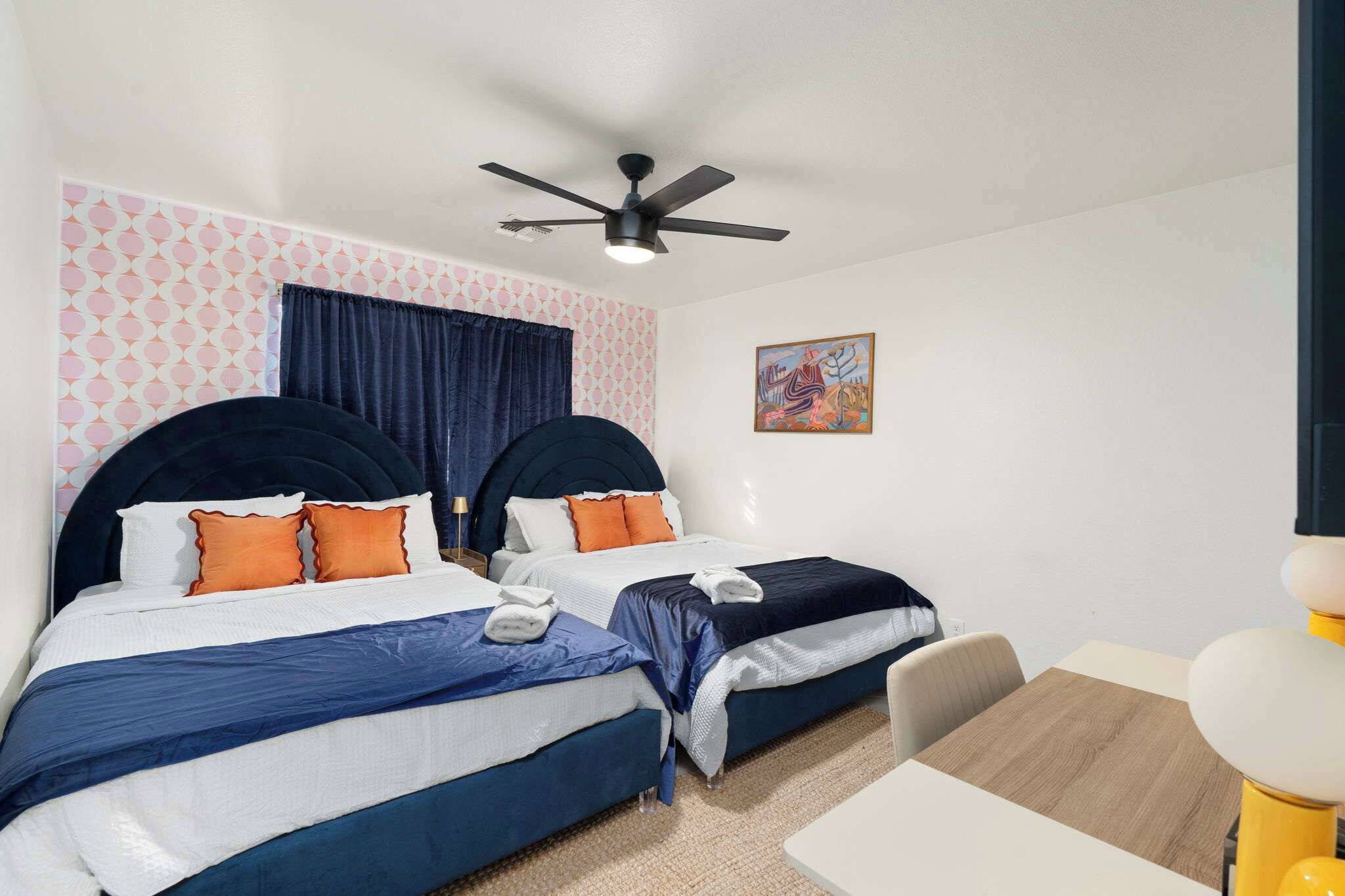 A hotel room with two beds, a ceiling fan, a painting on the wall, and a small table with a chair.