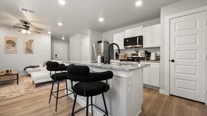 A modern kitchen with white cabinets, a stainless steel refrigerator, and a microwave. There is a bar area with two black stools and a granite countertop. - Wellness Retreat - 3BR/2BA - Family Friendly (Lubbock)