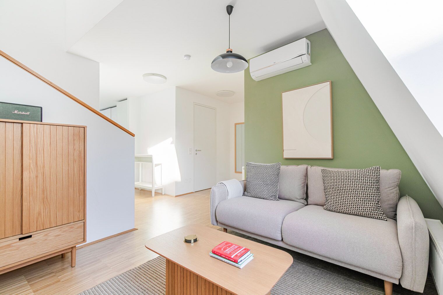 A modern living room with a wooden cabinet, a grey sofa with patterned cushions, a coffee table with books, and a staircase with a wooden railing.