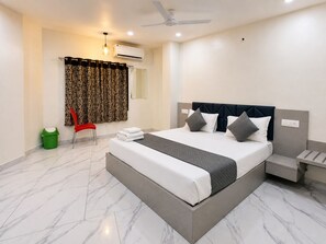 A hotel room with a bed, a nightstand, a chair, a trash bin, and a window with curtains. - Soneva Hotel (Ahmedabad)