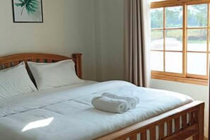 Una cama bien tendida con sábanas blancas y una toalla enrollada, un cabecero de madera y una ventana con cortinas.