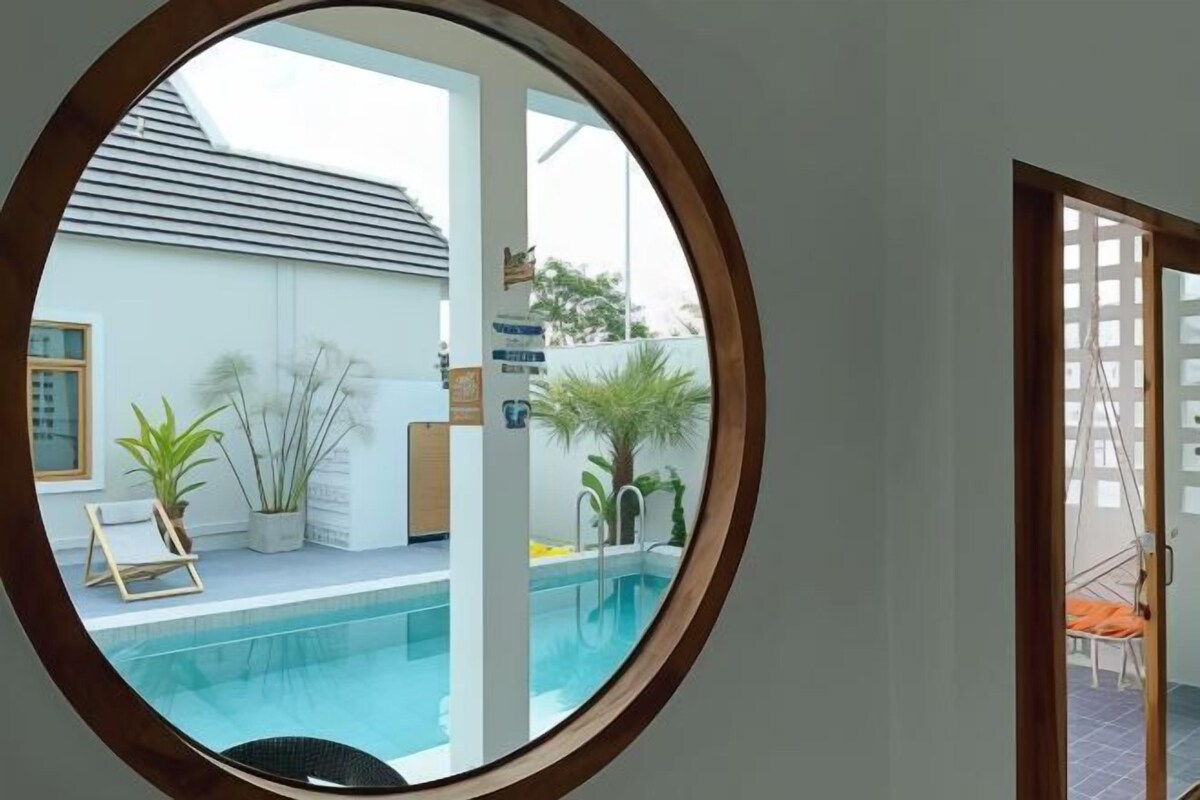Vista a través de un ventanal circular de una zona de piscina con un edificio blanco, plantas en macetas y una silla.