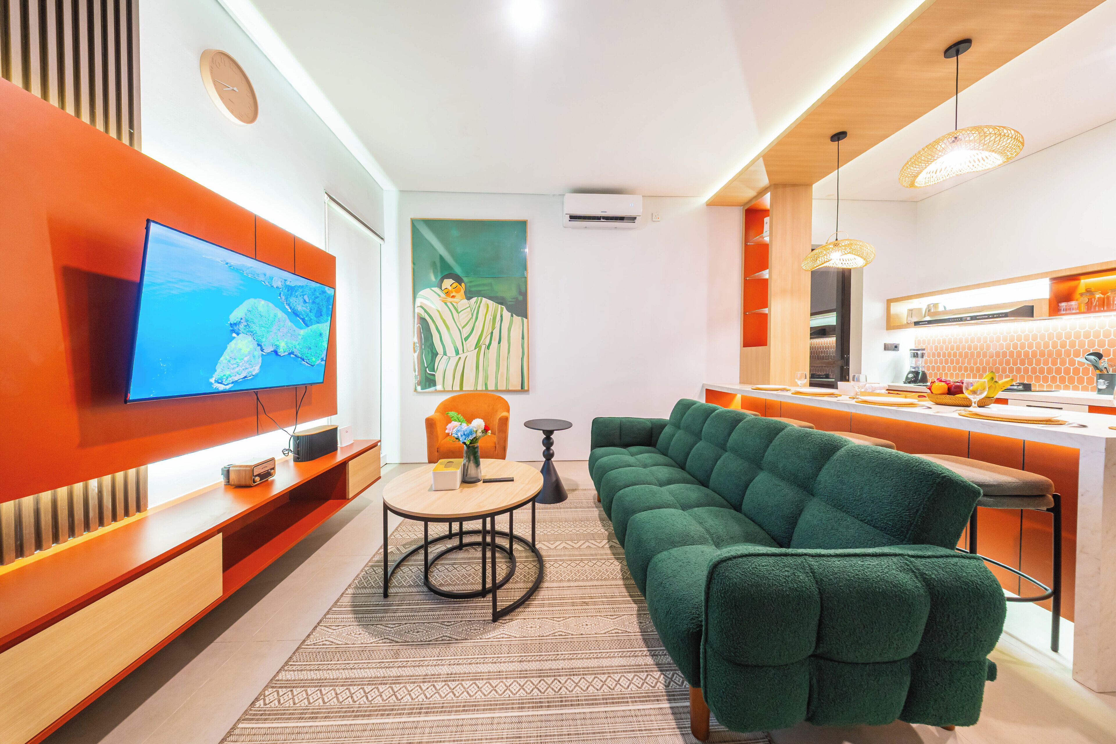 A modern living room with a flat-screen TV, a green sofa, a small round table, and a kitchen area with a bar.
