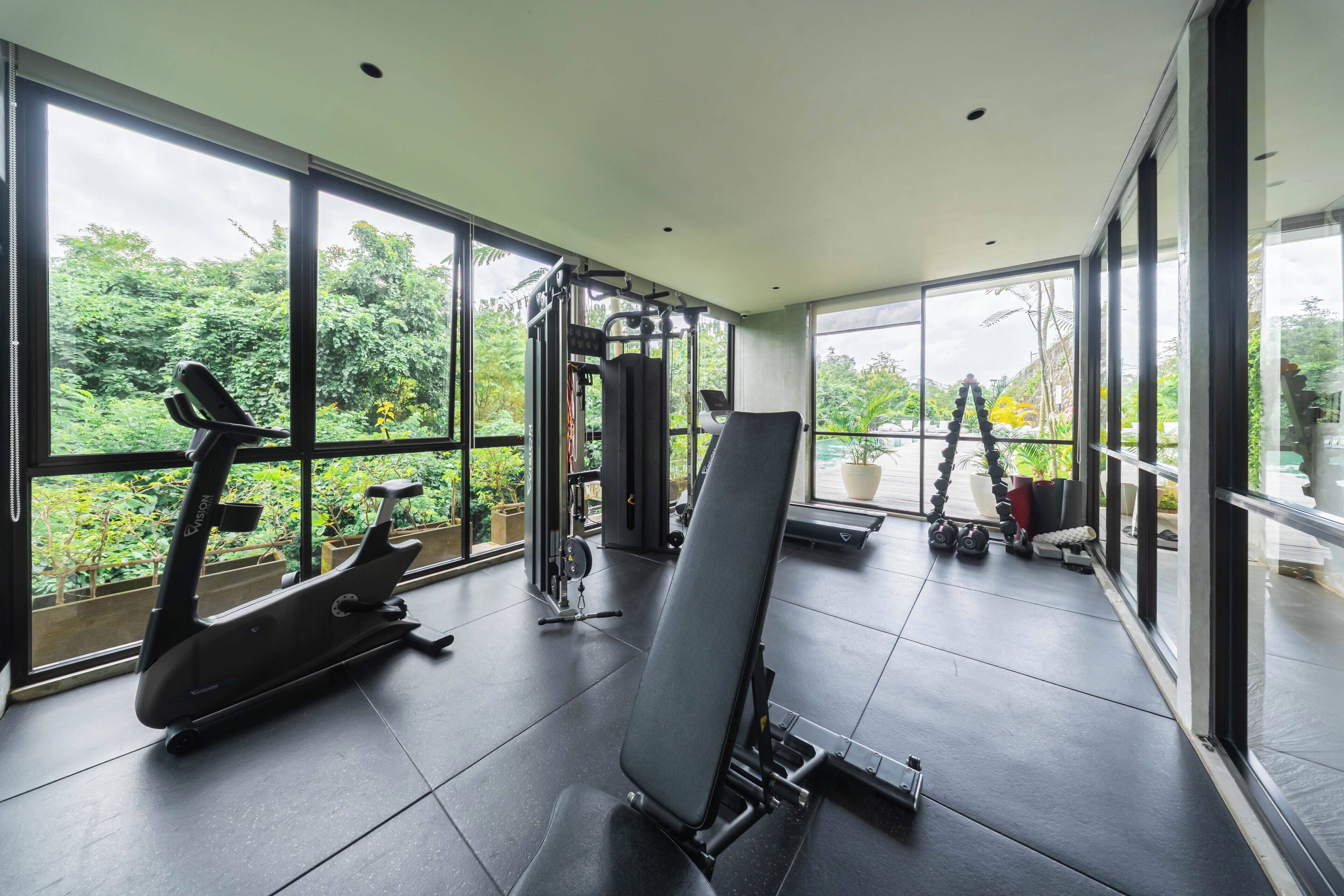 Un gimnasio con equipo de ejercicio, ventanas amplias y vistas a zonas verdes.