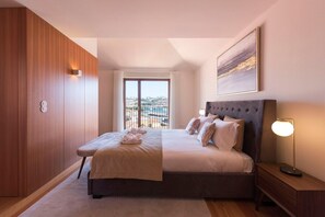 A modern hotel room with a large bed, a nightstand, a bedside lamp, a view of the city through a large window, and a wooden wardrobe. - Porto Riverviews | Monte dos Judeus (Porto)