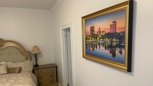 Un dormitorio con cama, mesita de noche, cómoda y un cuadro de un paisaje urbano enmarcado en la pared.