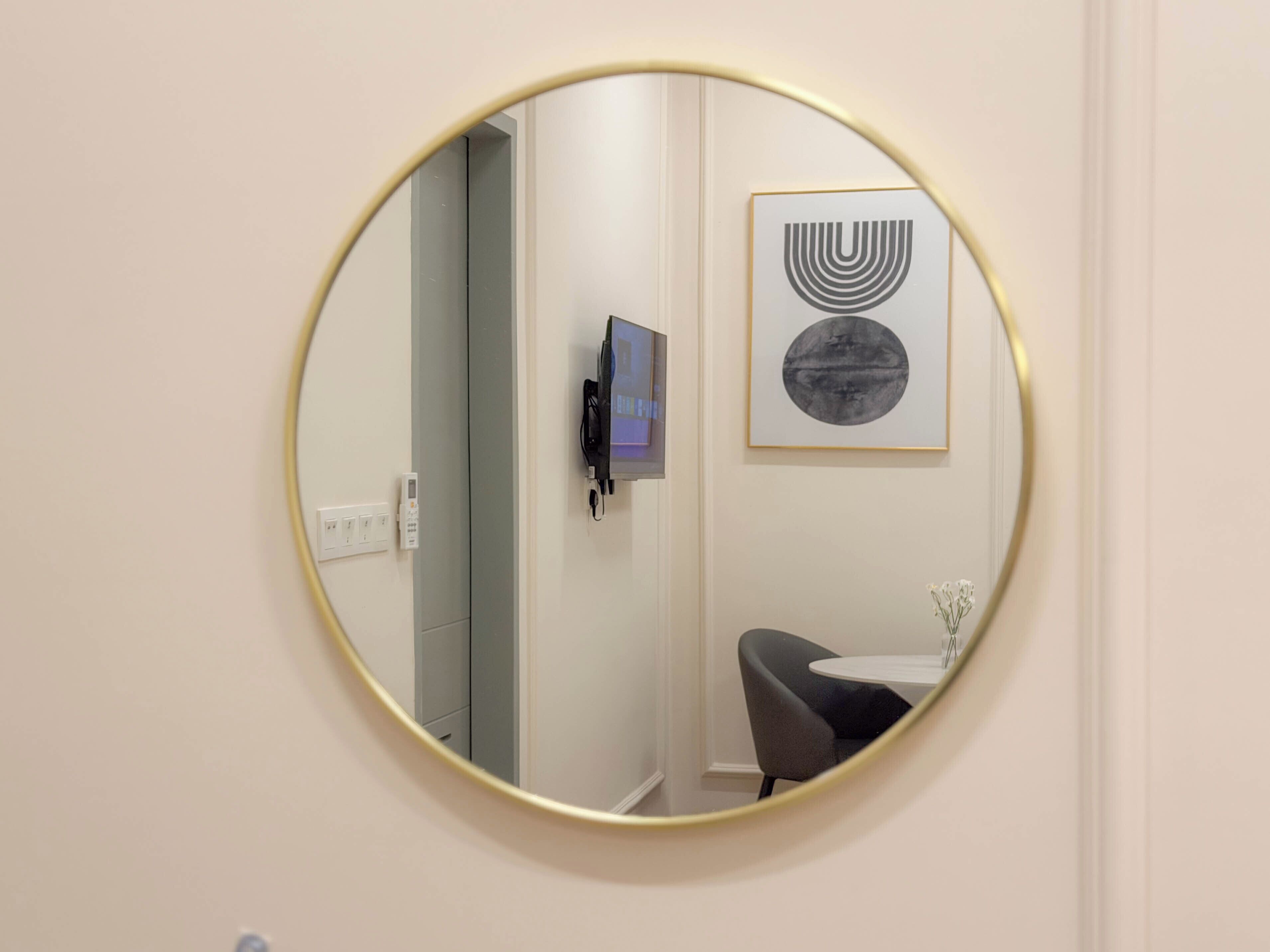 Un espejo redondo con marco dorado, un teléfono instalado en la pared, una obra de arte abstracta enmarcada y una mesita con un jarrón.