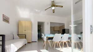 Une pièce moderne et bien éclairée, comprenant un lit, une armoire en bois, un coin repas avec des chaises et une table, et une cuisine à l’arrière-plan.