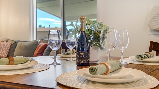 Una mesa de comedor dispuesta con platos, copas, una botella de vino espumoso Stella Rosa, un servilleta enrollada y una planta en maceta.